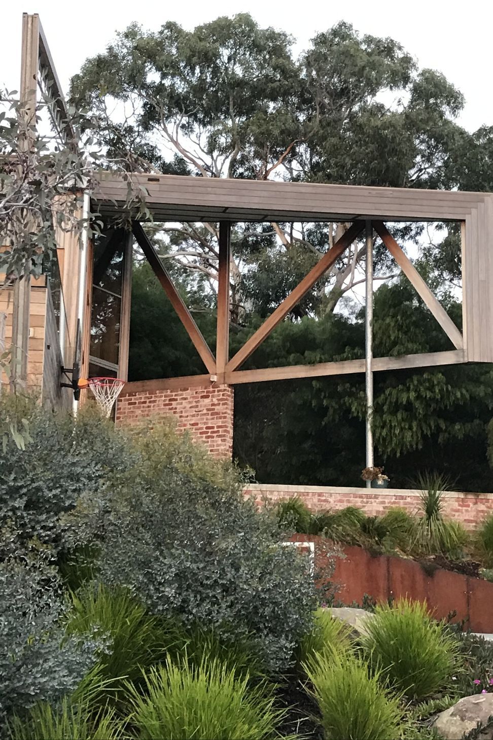 An exposed timber truss entry verandah roof hovers above a recycled brick wall, framing views of the native eucalypt canopy