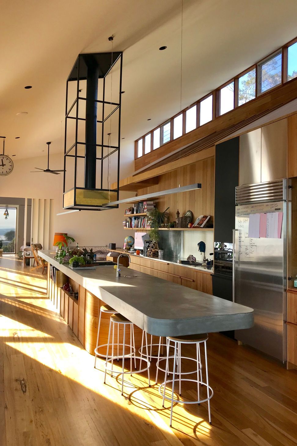 A custom industrial steel rangehood framework rises above a cantilever concrete island bench, beneath raked ceilings punctuated by high clerestory windows