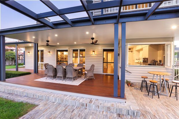 A covered outdoor entertaining area adjoining a contemporary weatherboard home. The space features a timber deck with a large outdoor dining setting, ceiling fans, integrated lighting, and a translucent roof pergola supported by dark structural posts. To the right is an open outdoor kitchen or servery with a benchtop, stools, and visible kitchen joinery beyond. Paved stone flooring leads up to the deck, while the house has horizontal light-coloured cladding and extensive openings.