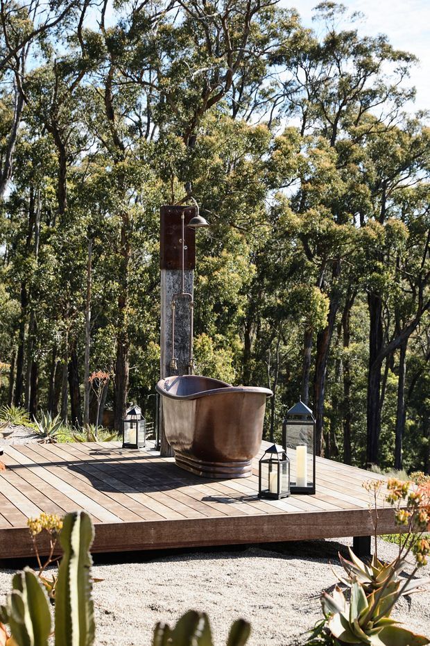 Outdoor freestanding copper-toned bathtub with an exposed shower fixture, positioned on a raised timber deck in a wooded natural setting. Decorative lanterns surround the bath, with gravel and drought-tolerant planting below the deck. The scene is sunlit and rustic, blending a spa-like bathing area with the surrounding bushland.