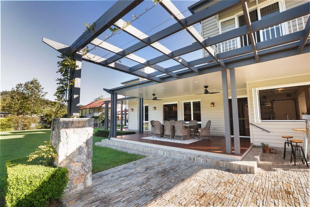 Exterior view of a light-coloured weatherboard home with a covered outdoor dining terrace, dark timber pergola, stone posts, paved courtyard and an open servery window. The setting includes landscaped lawn and hedging, with a second-storey portion of the house visible above.