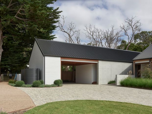 Exterior view of a contemporary single-storey residential building in a landscaped setting. The structure has a steep gable roof clad in dark corrugated metal, pale horizontal weatherboard cladding, a smooth light concrete wall, and a covered open carport or entry passage. A paved driveway and curved pedestrian path lead toward the building, with mature trees and planting surrounding the site.