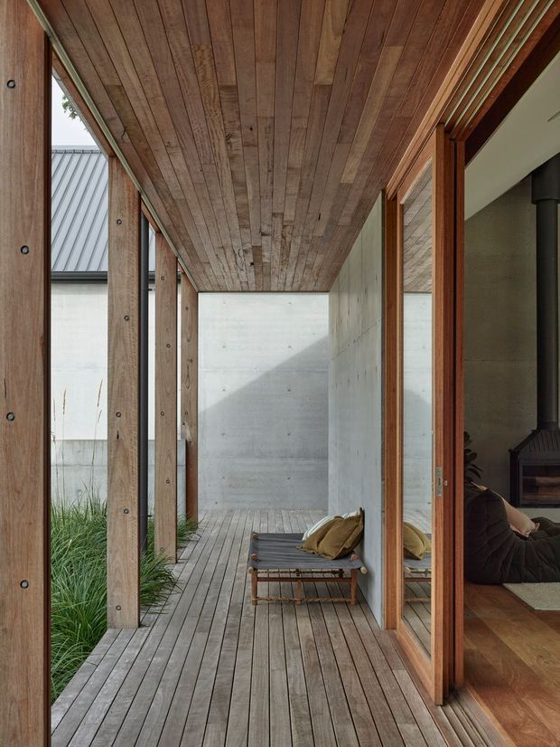 A covered timber deck or breezeway runs alongside a house, framed by substantial exposed timber posts and a timber-planked ceiling. Full-height timber-framed glazing opens to an interior living area with a dark freestanding stove, while a built-in timber daybed sits on the deck. Raw concrete walls, weathered timber decking, and planted grasses create a restrained indoor-outdoor composition with strong natural textures.