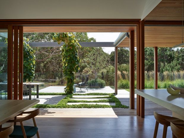 A warm contemporary interior opens directly onto a landscaped courtyard through expansive timber-framed sliding or folding glazing. The room includes a dining table and chairs at left and a white island or counter at right, with timber flooring, timber ceiling lining, and strong indoor–outdoor connection. Outside are paved paths, clipped groundcover, garden planting, and a pool or water feature area.