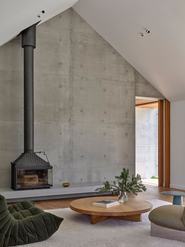 A refined living room with a dramatic exposed concrete feature wall and angled ceiling, centred around a suspended black wood-burning fireplace. The room includes a low timber coffee table, plush deep green seating, a rounded neutral armchair, warm timber door framing, recessed spotlights, and a large light rug. A glazed opening provides a view into another concrete-lined area, while indoor greenery and restrained furnishings create a calm contemporary atmosphere.