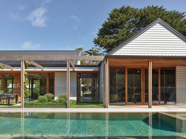 A single-storey residential house viewed from across a swimming pool. The home has a pitched gable-roofed wing with white horizontal weatherboard cladding, extensive timber-framed glazing and doors, and a connected low-pitched roof section. A timber pergola extends along the pool-facing facade, with landscaped planting and mature trees around the property.