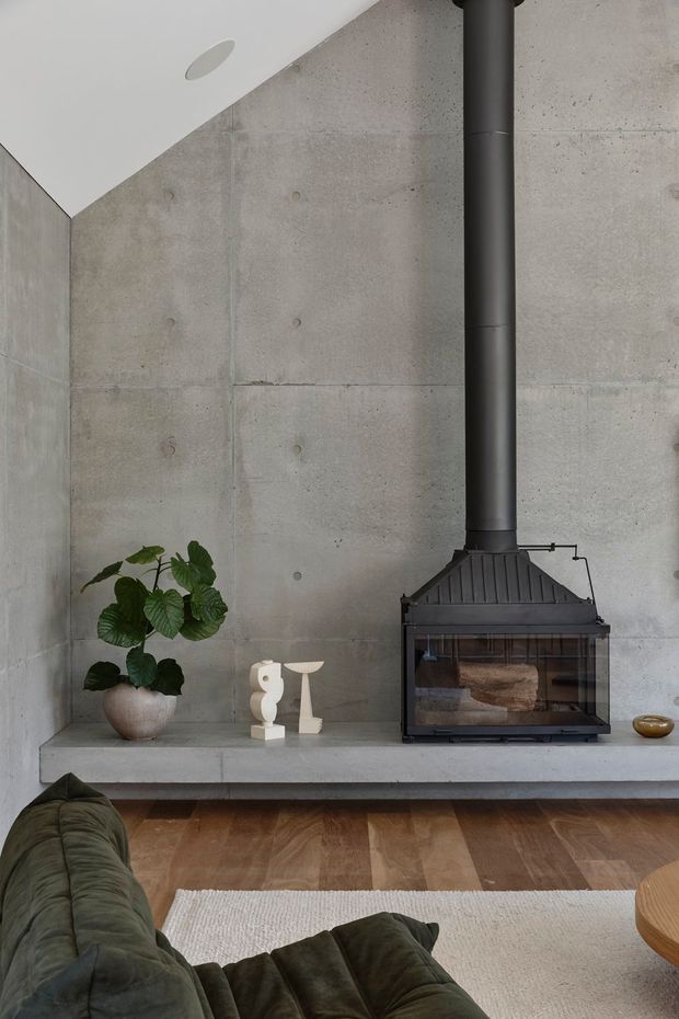 A minimalist living room focused on a black freestanding wood-burning fireplace set against a raw board-formed concrete feature wall. The fireplace sits on a substantial concrete hearth with a small plant and sculptural decor, while a dark green sofa, timber floor, and pale rug are visible in the foreground. The space has a restrained, contemporary industrial character with clean lines and muted natural tones.