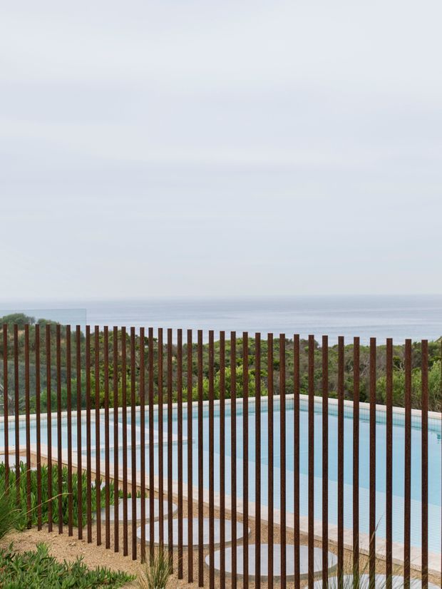 A contemporary outdoor swimming pool overlooking a calm ocean horizon. Tall, slender rust-coloured vertical metal posts form a transparent privacy and safety screen in the foreground, with pale stone paving, low coastal planting, and a curved pool edge visible behind it. The scene is softly lit and muted, creating a serene coastal atmosphere.