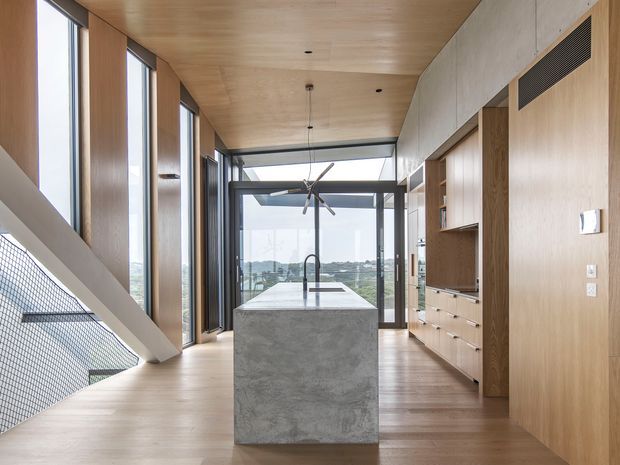 A contemporary open-plan kitchen with a monolithic grey concrete island and integrated sink, surrounded by light timber cabinetry and matching timber wall and ceiling finishes. Full-height glazing opens to a distant suburban view, while a stair with mesh balustrade is visible on the left. Dark metal window frames and a sculptural pendant light add contrast.