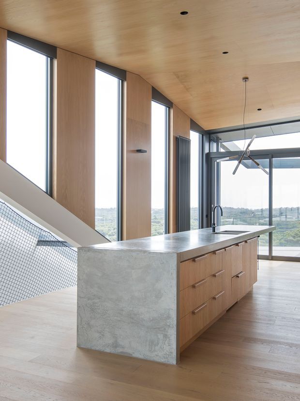 A bright, open-plan contemporary kitchen centred on a long island with a pale concrete waterfall end and timber drawer cabinetry. Full-height glazing provides expansive views, while vertical timber wall panels, a matching timber ceiling, recessed downlights, and a modern pendant reinforce the clean architectural design. A stair with mesh balustrade is visible at left.