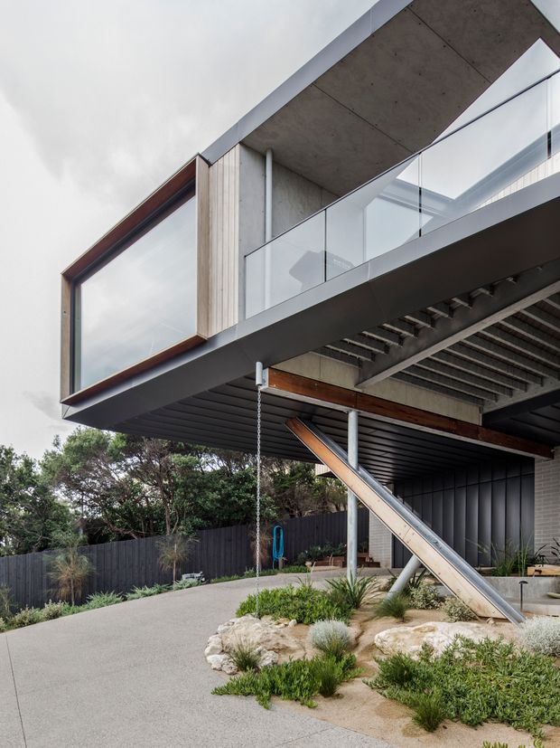 A contemporary elevated house exterior with a dramatic cantilevered upper floor, exposed concrete soffit, timber-framed corner glazing, clear glass balustrades, dark metal cladding, and exposed structural steel supports. The landscaped foreground includes low native-style planting, rocks, and a concrete driveway or outdoor approach.