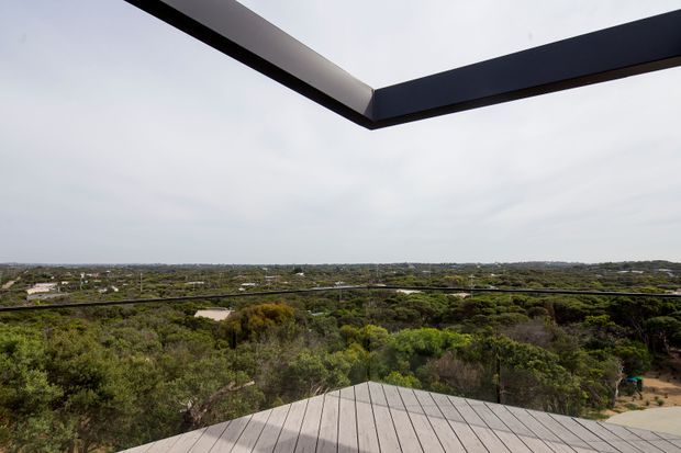 A wide elevated outdoor view across dense coastal-looking vegetation and a low suburban landscape, framed by a dark angular roof or canopy edge above and a pale timber deck in the foreground. The image emphasises the open outlook and architectural framing rather than a specific enclosed room.