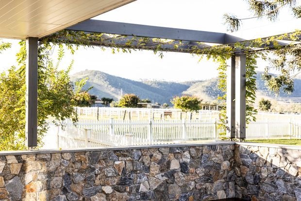 Covered outdoor terrace or patio viewed from beneath a flat pergola-like roof, with dark structural beams, climbing greenery, and a substantial rough-stone retaining or boundary wall. A white picket fence and landscaped grounds are visible beyond, with wooded mountains in the distance. The image has a bright, warm outdoor atmosphere and highlights the sheltered entertaining area and its mountain setting.
