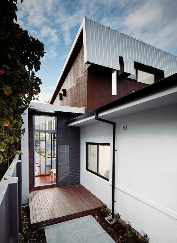 Exterior view of a compact contemporary multi-level residence surrounding a small timber-decked courtyard or entry terrace. The facade combines dark brick, vertical timber cladding and crisp white walls, with a strongly angled gable roof clad in corrugated metal. Black-framed glazing and a glazed connecting section open toward the deck; a dog is visible inside. The image is photographed from an outdoor walkway beneath a partly cloudy sky.