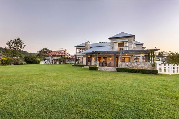 Exterior view of a substantial multi-level residential house set within a broad manicured lawn. The home has a complex pitched and hipped roofline, pale horizontal weatherboard-style cladding, extensive covered verandah and balcony areas, timber-toned posts and trim, and a stone-clad lower facade. A white picket fence and a separate pavilion-like structure with a red metal roof are visible on the property.