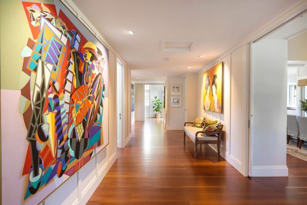 A bright, refined residential hallway with polished dark timber flooring, white painted walls and detailed traditional trim. Large colourful abstract artworks line the corridor, with a small upholstered bench and console-style seating area along the right wall. Doorways open to adjoining rooms, including a dining area, while recessed ceiling lights and an airy sightline emphasize the length of the space.