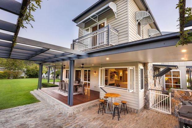 Exterior view of a multi-level weatherboard house with a broad covered outdoor entertaining area, upper-level balconies, white decorative railings, and a stone-clad lower section. The home opens onto a paved terrace with outdoor dining and bar seating, while a landscaped lawn and pergola-like roof structure extend to the side.