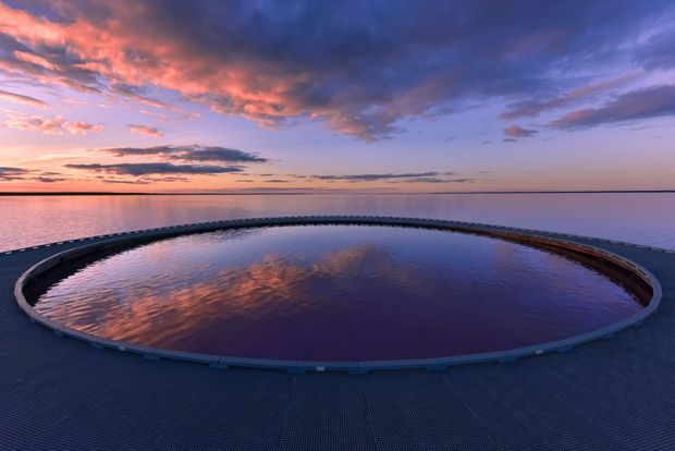 A circular infinity-edge pool or water feature viewed at sunset, surrounded by a dark deck and blending visually into a calm body of water on the horizon. The scene is dominated by pink, purple, and blue reflections beneath a dramatic cloud-filled sky.