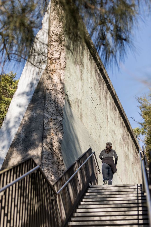 Vertical outdoor architectural photograph looking up a broad flight of concrete stairs with black metal handrails beside a very tall, weathered pale concrete wall. A person is walking uphill, while trees and blue sky frame the structure. The image emphasizes the monumental scale and textured concrete construction.