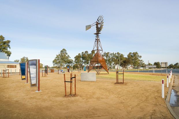 A broad outdoor recreational or civic landscape with a sandy, sparsely planted open area, young supported trees, a tall vintage windmill structure, and a freestanding sign or interpretive panel. Low-rise buildings, fencing, paved paths, and sporting or community infrastructure are visible in the background under a clear sky.