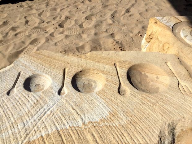 Close-up of a sandy beach sculpture or sand artwork featuring carved shell-like forms and elongated grooves, with textured sand surrounding the raised relief.