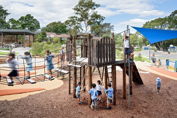 Outdoor children's playground with timber climbing structures, rope bridges, slides, and play equipment beneath a large blue shade sail. Several children are using the equipment, with landscaped grounds and low buildings in the background.