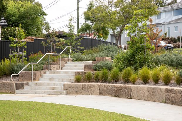 A landscaped suburban front garden with broad concrete steps and stainless-steel handrails leading up through terraced planting beds. Rough stone retaining walls, ornamental grasses, shrubs, mature trees, and a dark privacy fence define the garden, with a light-coloured multi-storey house visible behind.