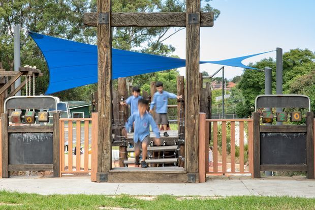 Outdoor recreational play area with a rustic timber-framed entry and stair-like play structure, enclosed by timber railings and protected by bright blue triangular shade sails. Children are using the structure, with landscaped greenery and residential surroundings in the background.
