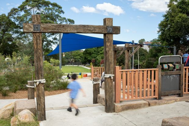 Outdoor landscaped recreational area with a rustic timber entry portal, concrete paths, timber safety railings, large blue shade sails, planting, and a child moving through the entrance. The setting appears to be a playground or public play space.