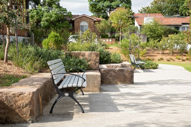 A landscaped outdoor pedestrian space with concrete paving, rustic stone retaining walls and built-in seating, surrounded by mature planting and suburban brick homes. Dark metal-and-timber benches line the walkway, creating a shaded garden-like public or residential communal setting.