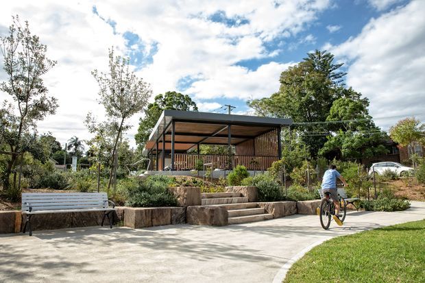 A landscaped outdoor public or residential garden setting with a small contemporary pavilion featuring a flat roof, open sides, timber screening, and a covered seating area. Broad concrete paths, terraced stone retaining walls, planted beds, a bench, and a cyclist are visible beneath a bright partly cloudy sky.