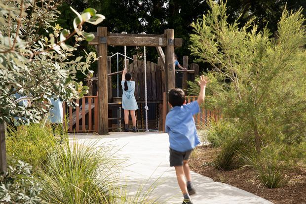 A landscaped outdoor pathway leads toward a rustic timber gateway and fenced play structure. Children are visible playing near the entrance, surrounded by ornamental grasses, shrubs, and mature greenery. The scene appears to be a designed recreational or outdoor play area.