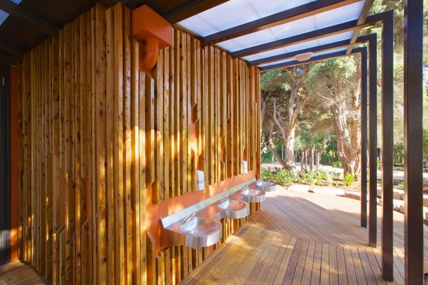 A covered outdoor deck or verandah with a warm timber-slatted wall, built-in orange-toned bench, and several small round stainless-steel basins or fixtures. A translucent roof is supported by dark metal framing, while timber decking extends toward a landscaped, tree-filled outdoor setting. The design has a strong contemporary architectural character with warm natural materials and open-air connectivity.
