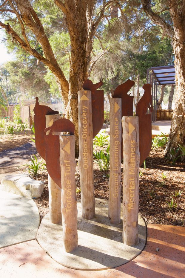 Outdoor educational garden or nature-play area beneath mature trees, featuring sculptural corten-steel animal silhouettes mounted alongside rough timber signposts. The posts carry vertical lettering such as “Outdoor Discovery,” “Ground Learning Space,” and “Meer... Matters,” with a paved walkway, planted garden beds, and a covered timber structure in the background. Bright, natural, rustic setting.