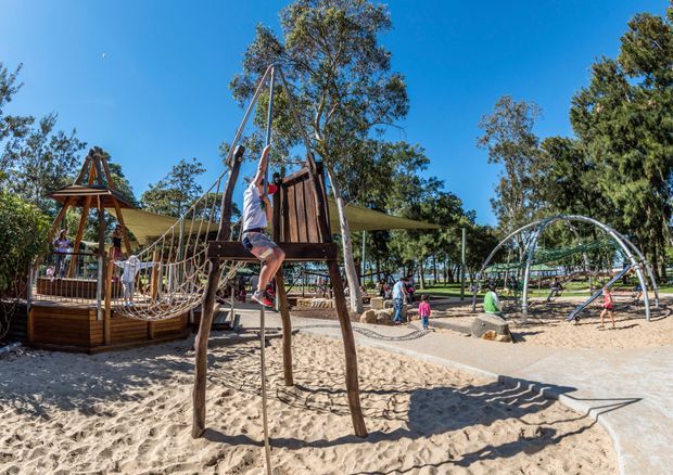 A sunny outdoor playground with extensive sand surfacing, timber climbing structures, rope nets, shade sails, and children playing. The scene includes a prominent timber-and-rope climbing frame in the foreground, additional play equipment, paved paths, mature trees, and open sky.