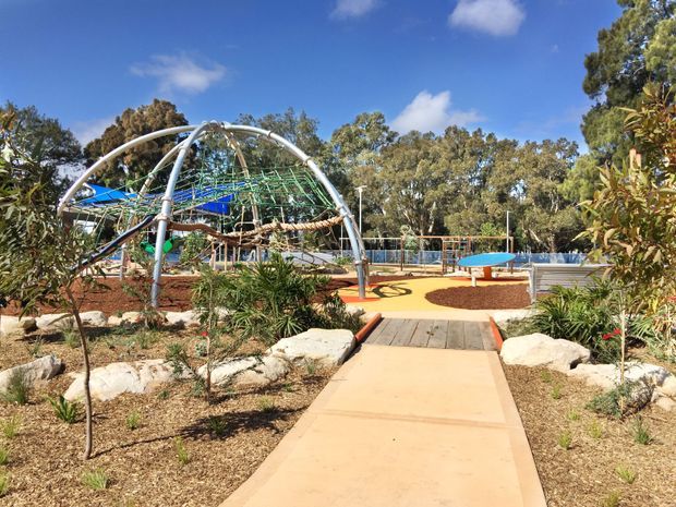 A landscaped outdoor recreational area with a concrete pathway leading over a small timber footbridge toward a colourful playground and climbing structure. The site includes native-style plantings, rocks, timber edging, and mature trees under a bright blue sky.