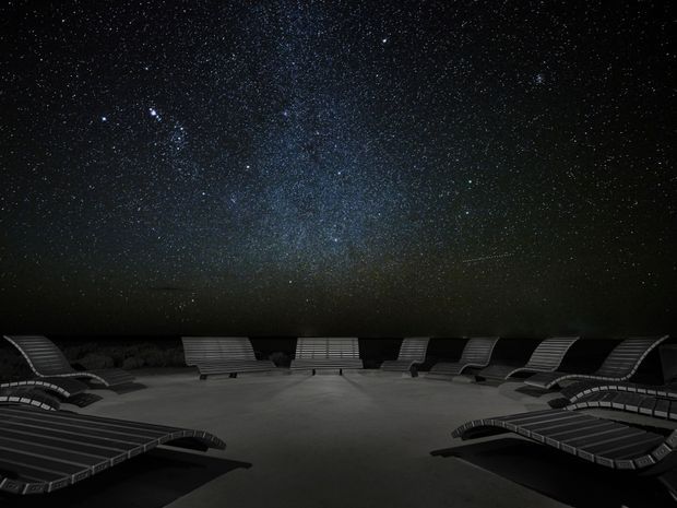 A dark outdoor terrace or poolside area arranged with numerous curved lounge chairs in a circular layout beneath a dramatic star-filled night sky, including a visible Milky Way. The scene is monochromatic and atmospheric, with the lounge furniture silhouetted against the open landscape.