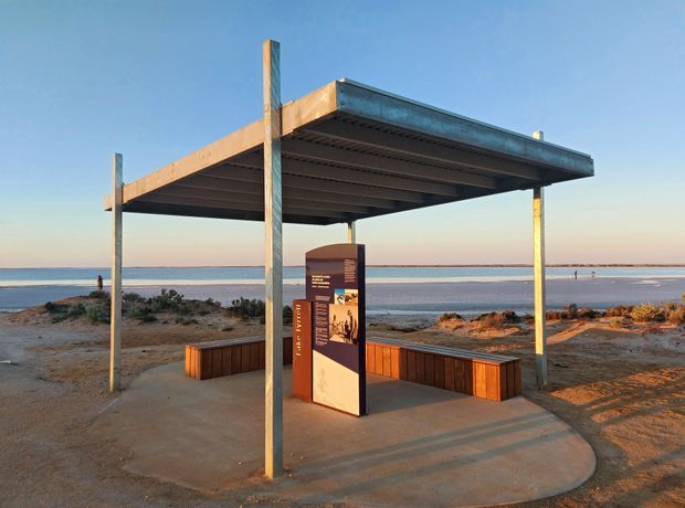 A modern open-sided beach viewing shelter with a flat metal roof supported by slender steel posts. Built-in timber bench seating surrounds an interpretive information panel on a concrete pad, overlooking a calm shoreline and shallow water. The setting is sparse, coastal, and strongly defined by the shelter's simple contemporary construction.