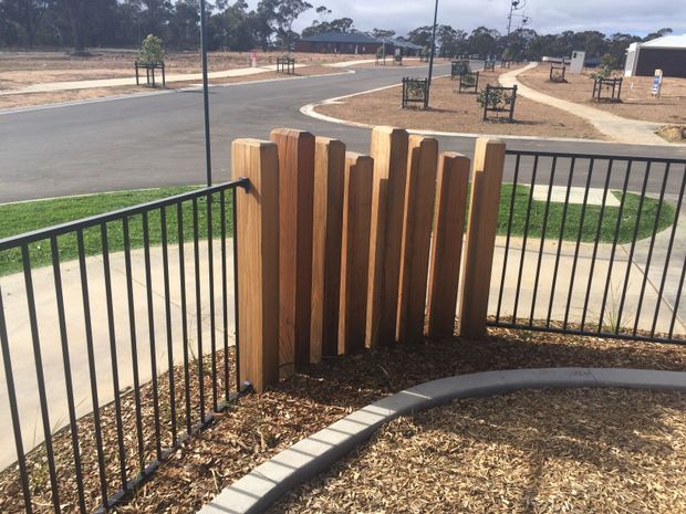 A landscaped residential streetscape viewed from a raised outdoor area, featuring black metal balustrade fencing, a curved concrete garden edging, mulch, and a decorative vertical timber privacy screen. A paved footpath, roadway, newly planted trees, and modern homes are visible beyond.