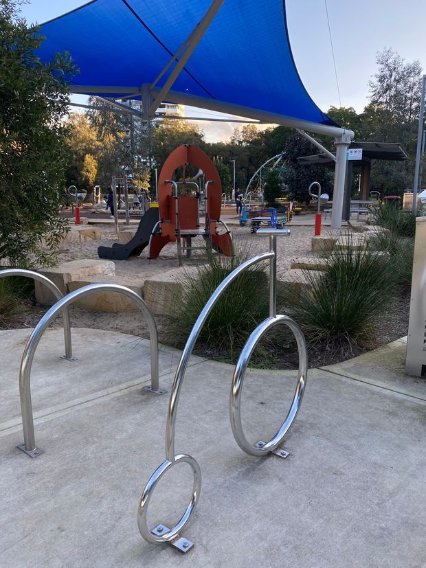 Outdoor public playground beneath a large blue tensile shade canopy, with stainless-steel looping bike racks in the foreground, concrete paving, landscaped plantings, playground equipment, and seating or shelter structures in the background.