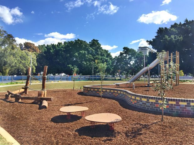 A landscaped outdoor playground with timber climbing and balancing structures, spring-mounted round platforms, a metal slide, young trees, and a curved low retaining wall faced with multicoloured brick tiles. The play area is covered in brown wood mulch and enclosed by fencing, with mature trees and a bright blue sky in the background.