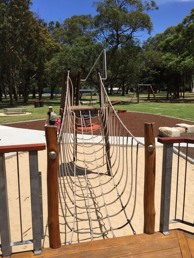 A rope climbing bridge and play structure in a landscaped public park, with timber posts, metal railings, a sandy play surface, grass, mature trees, and additional playground equipment in the background.