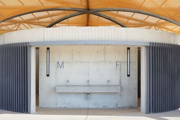A symmetrical semi-enclosed washroom or utility facility beneath a large curved, exposed steel-framed canopy. The central concrete wall has two narrow vertical mirrors, multiple wall-mounted taps, and a long concrete trough basin with a central outlet. Dark corrugated metal side screens and a pale corrugated upper enclosure contrast with the warm translucent roof structure. The space is utilitarian, industrial, and minimally detailed.