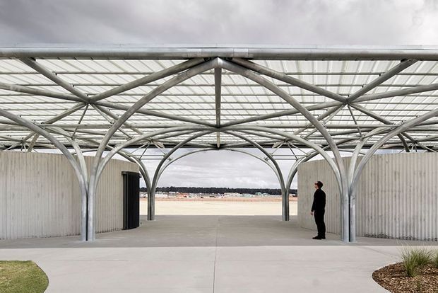 Open-air architectural pavilion with a translucent roof supported by an expressive network of curved and branching steel columns and beams. Corrugated pale walls frame a broad central opening toward a sandy waterfront or coastal landscape, with a concrete forecourt and sparse planting. A person provides scale beneath the large lightweight canopy.