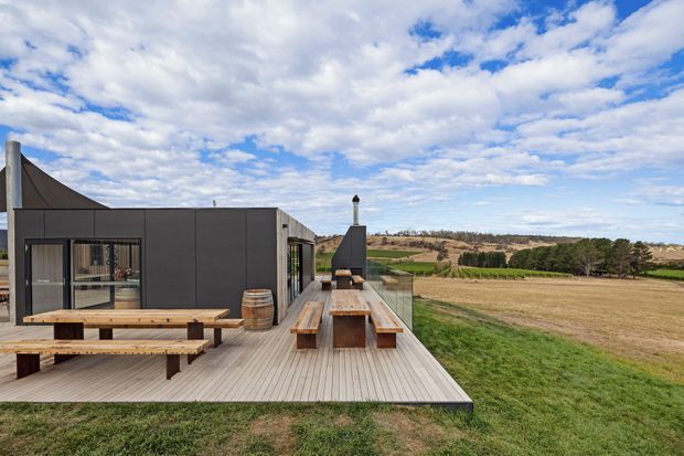A contemporary rural home with dark, rectilinear cladding opens onto a broad timber deck overlooking expansive farmland. The deck includes long built-in timber dining tables and benches, a tall outdoor fireplace or chimney structure, and clear glass balustrading. Rolling cultivated fields, scattered trees, and a partly cloudy blue sky form the distant setting.