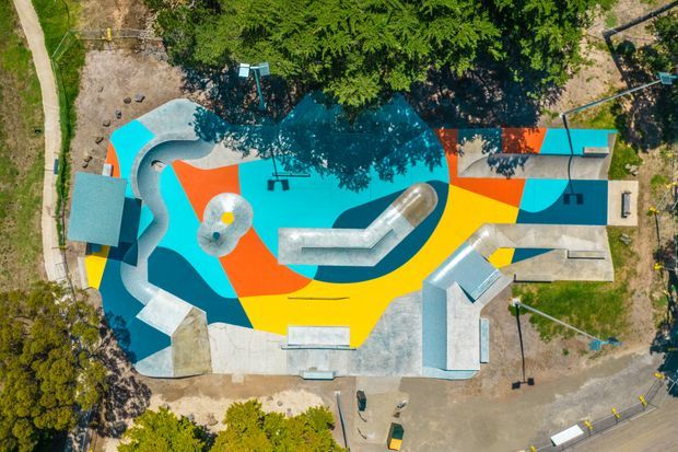 Aerial view of a brightly painted outdoor skate park with interconnected concrete ramps, bowls, rails, and covered structures. The park features bold turquoise, orange, yellow, dark blue, and red geometric surface graphics, surrounded by trees, paths, and open ground.