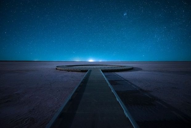 A symmetrical nighttime view along a narrow linear walkway or pier leading to a circular platform set across a broad, dark landscape. The horizon glows beneath a deep blue, star-filled sky, creating a minimalist and atmospheric scene.