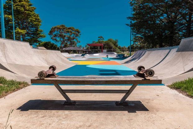 A brightly coloured outdoor skate park viewed from the end of a concrete half-pipe, with a weathered wooden grind rail and skateboard wheels in the foreground. The park includes curved concrete ramps, blue and orange painted riding surfaces, mature trees, and clear sunny skies.