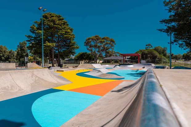 Outdoor concrete skate park with interconnected bowls, ramps, and smooth transitions, featuring a bold geometric mural in turquoise, yellow, orange, and blue across the floor. Mature trees, tall lighting poles, and a shaded structure surround the recreational facility under a clear sky.