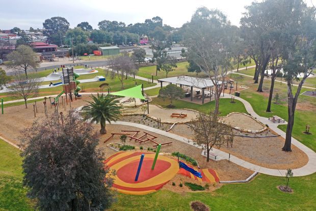 Aerial view of a landscaped public playground and recreation area, with colourful play equipment, shade sails, winding paved paths, picnic tables, mature trees, lawns, and a covered pavilion. Residential and street infrastructure are visible in the background.